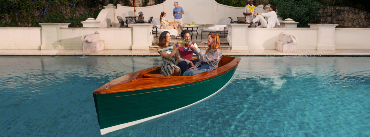 People enjoying a leisurely boat ride in a clear blue pool or canal, surrounded by modern architecture.