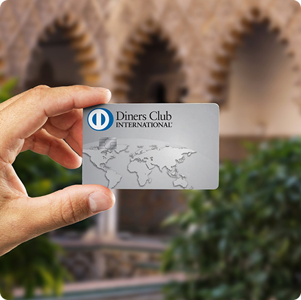 Hand holding up a silver loyalty or membership card with a circular blue logo against a blurred architectural background.